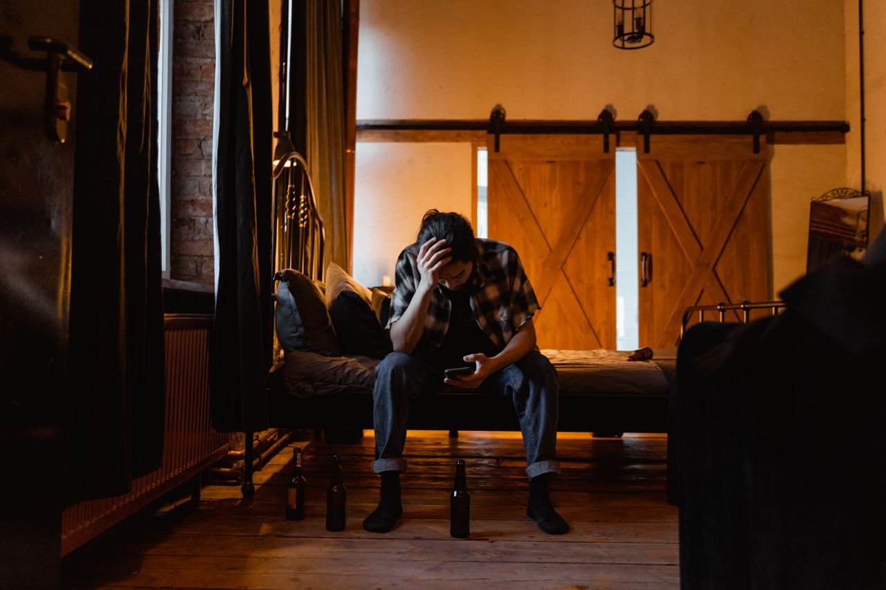heros-img A man sits on a bed in a dark room, appearing pensive and stressed.